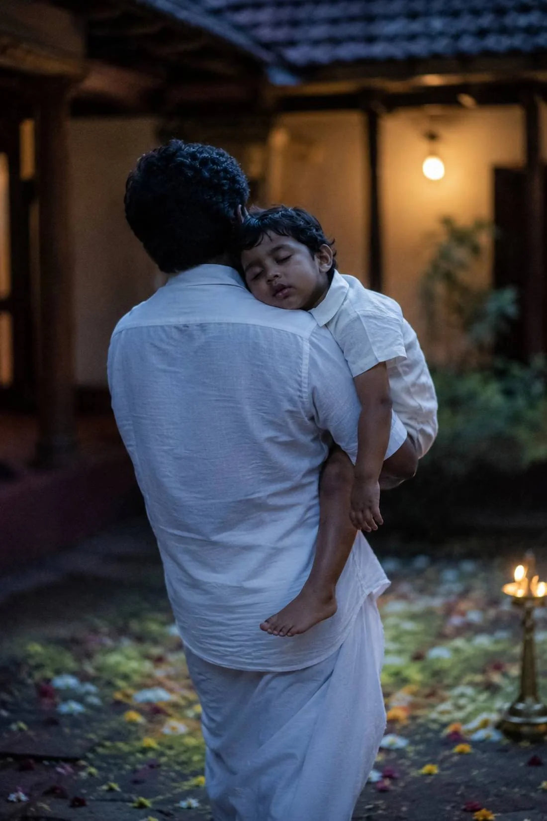 A father in a mundu carrying his sleeping child past the lamps at the end of Onam