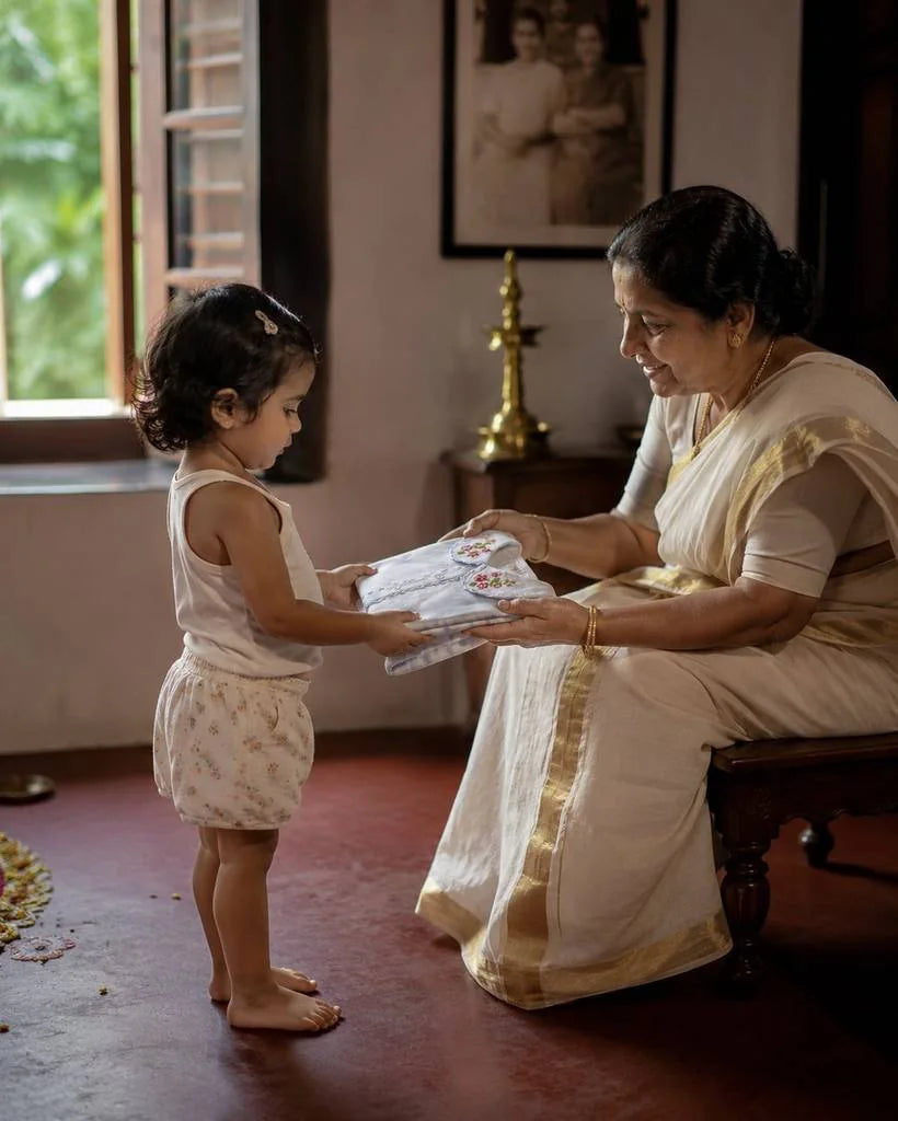 A grandmother in a kasavu saree handing folded new clothes to her granddaughter