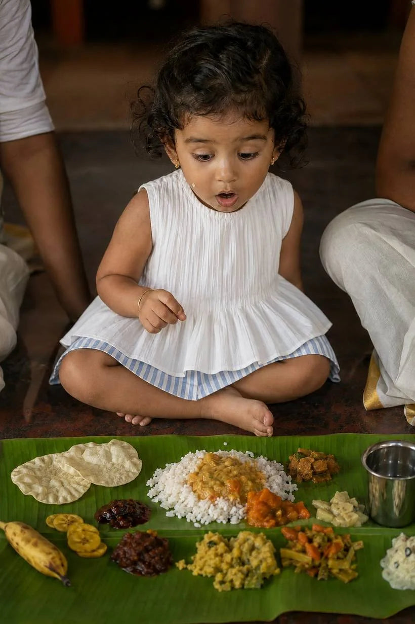 A toddler in white organic cotton sitting before an Onam sadya on a banana leaf