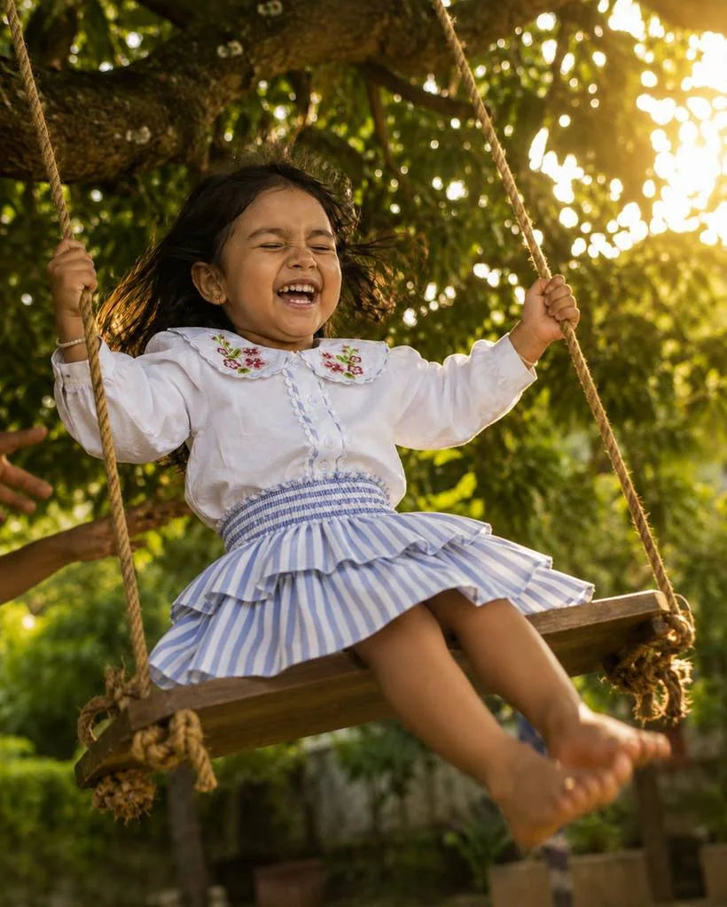 A little girl laughing on a swing in an organic cotton blouse and striped skirt